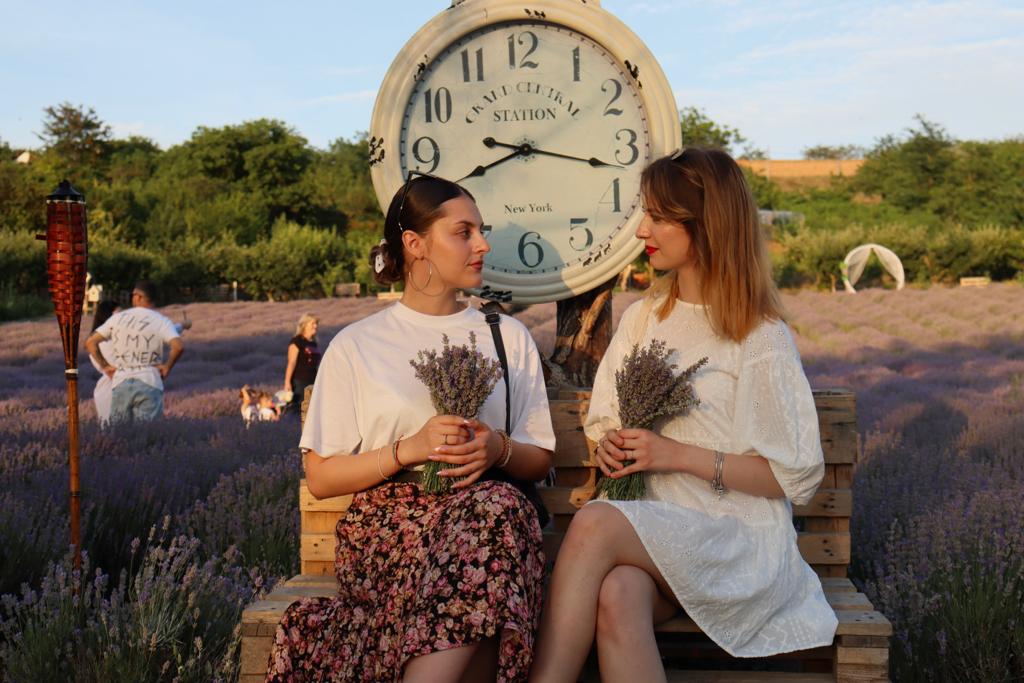2 fete așezate pe o bancă, având un ceas mare în spatele lor
